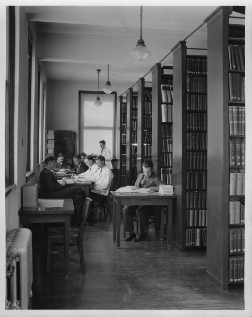 A black and white photo showing students in suits studying and bookshleves