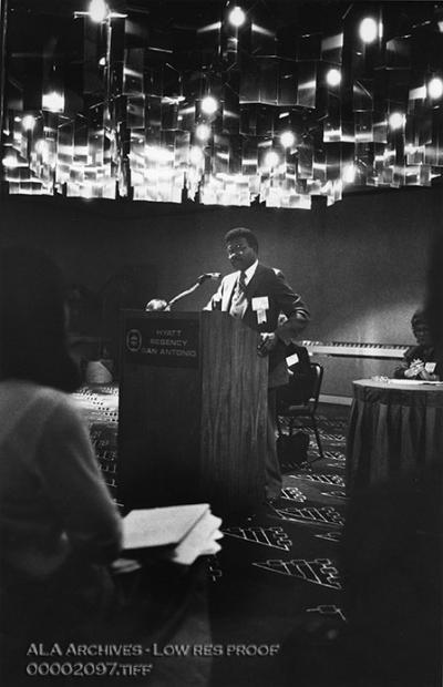In a darkened room, an African-American man speaks to an audience.