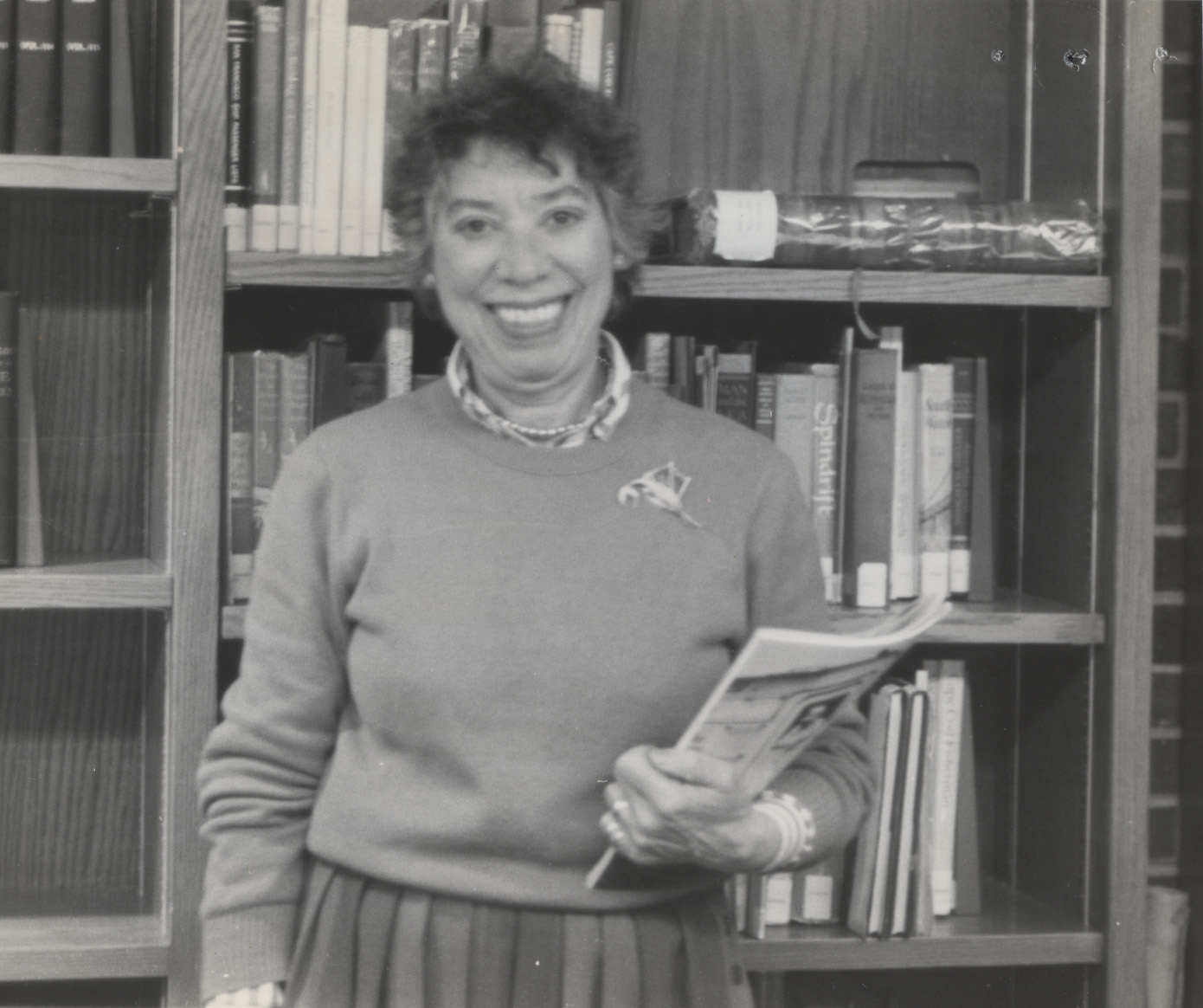 a black and white photo of a woman against a background of books, she is holding a periodical